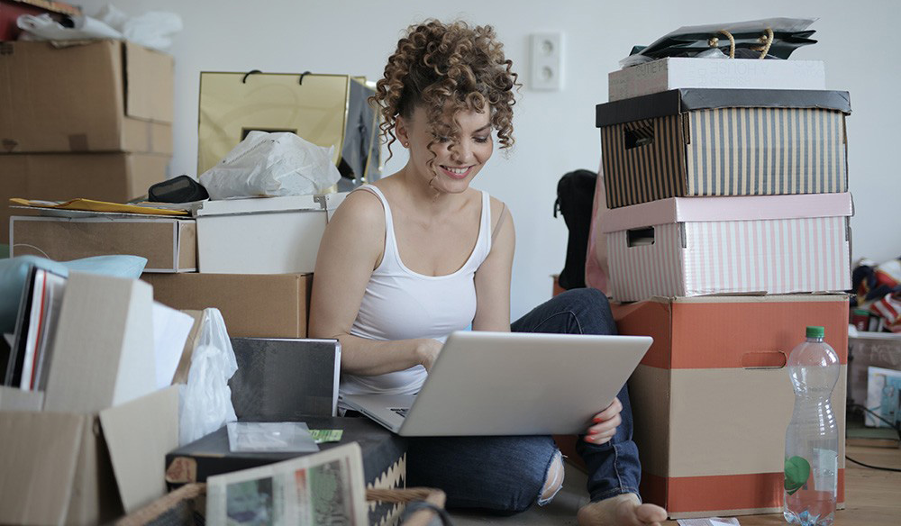 A young woman with curly hair sits on the floor amidst packed boxes, and she is looking into an open laptop.