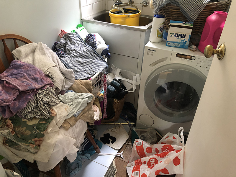 A messy laundry contains piles of clothes, a distant sink with a bucket in it, and a washing machine