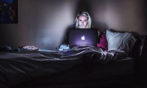A woman in the dark is illuminated by her Macbook screen as she sits in bed musing things people say on Facebook