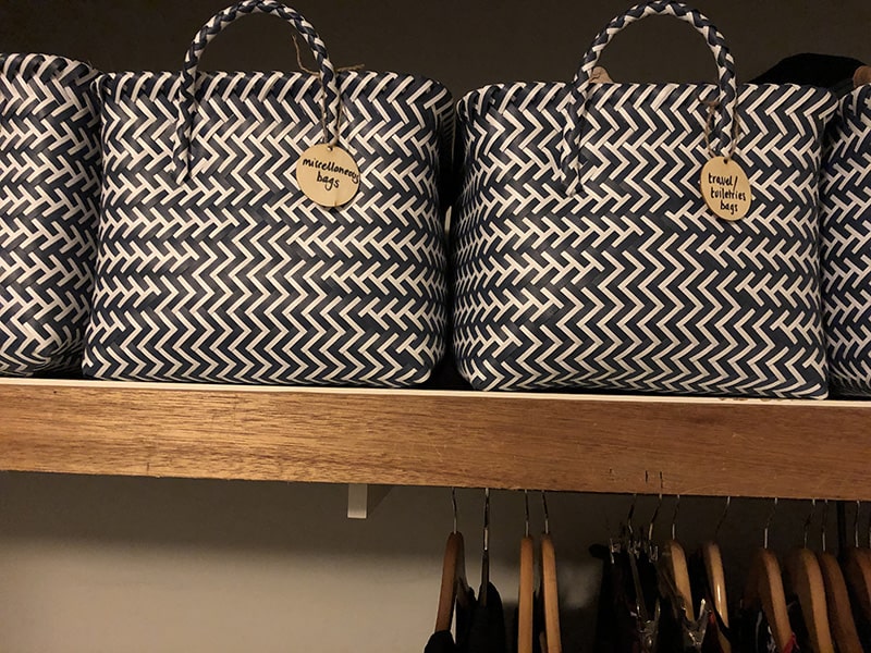 4 black and white baskets with swing tags sit on a shelf above hanging clothes. Label your stuff