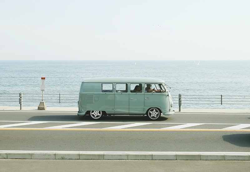 A pale blue Volkswagon combi van travelling with family on a road with an ocean backdrop