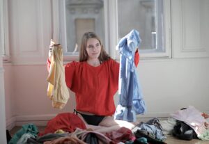 A young woman sits on the floor amidst clothes holds up a garment in each hand to declutter her clothes