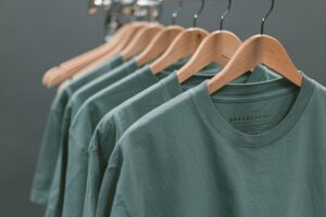 How many T shirts? 5 identical green T shirts are lined up on wooden coat hangers