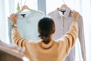 The rear view of a woman shows her holding up a knit sweater in each hand, decluttering and making decisions